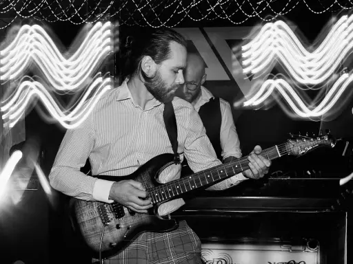 Muzyk gra na gitarze elektrycznej, a za nim stoi drugi mężczyzna. Efekt fal świetlnych sugeruje dynamiczną ekspozycję co to za moment.