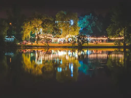 Nastrojowa fotografia nocna nad jeziorem, z oświetlonymi drzewami i budynkami, tworzącymi piękne odbicia w wodzie.