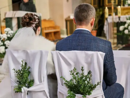Para młoda siedzi w kościele. Krzesła ozdobione są zielonymi gałązkami. Kto może być świadkiem na ślubie?