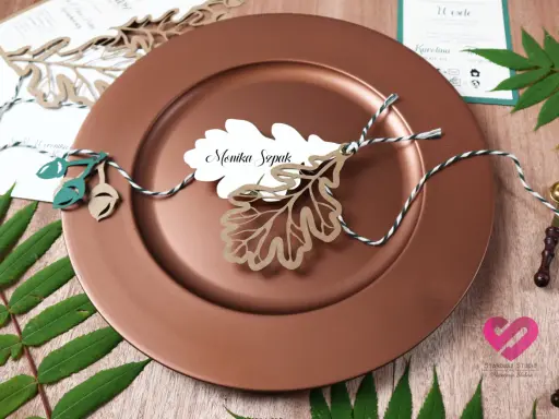 Winietki co to? Eleganckie winietki z imieniem "Monika Szpak" na liściu dębu, ozdobione sznurkiem i żołędziami, na brązowym talerzu.