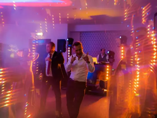 Wesołe uroczystości weselne inaczej! Dwóch mężczyzn tańczy w świetle dyskotekowej kuli i kolorowych świateł, DJ gra muzykę w tle.