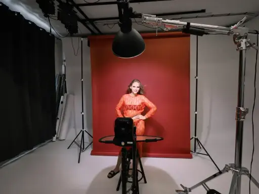 Oświetlenie studyjne przykłady: modelka w pomarańczowej sukience pozuje na tle czerwonego tła, otoczona sprzętem fotograficznym i lampami.