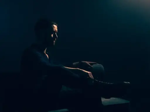 Mężczyzna w ciemnym pomieszczeniu, siedzi w fotelu. Jego sylwetka jest ledwo widoczna, podkreślona przez subtelne światło. To idealny przykład low key fotografii.