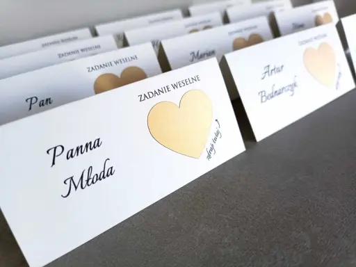 Białe karty z imionami gości i złotymi sercami, zapowiadające śmieszne zadania dla gości weselnych.