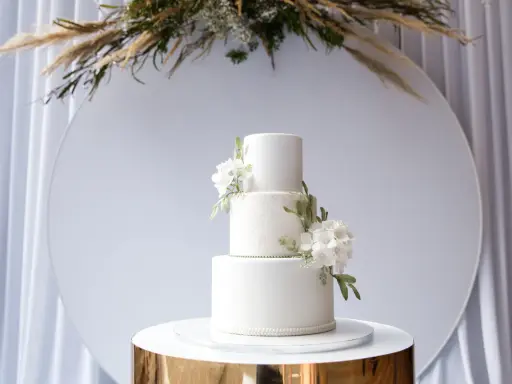 Elegancki, trójpoziomowy tort weselny w nowoczesnym stylu, ozdobiony białymi kwiatami i zielonymi liśćmi. Idealny na wyjątkowe okazje.