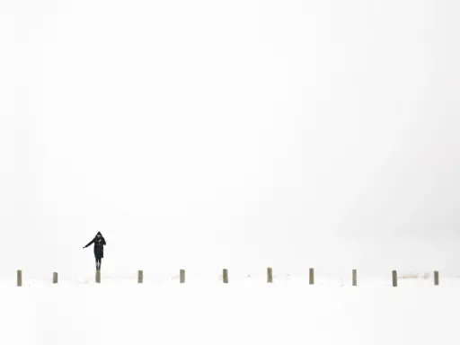 Osoba w czarnym płaszczu balansuje na słupku w śnieżnym krajobrazie. Minimalistyczna, high key fotografia.