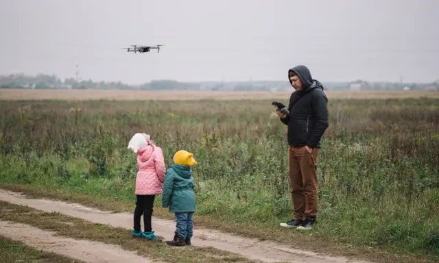 Ojciec steruje dronem, a dzieci z zaciekawieniem patrzą w niebo. Rodzinna przygoda z technologią w tle jesiennego krajobrazu.