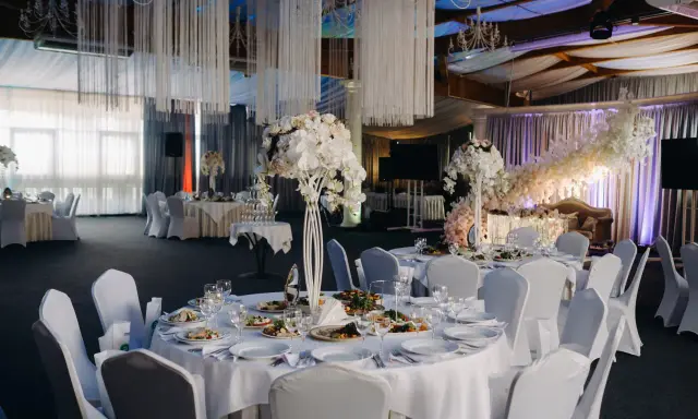 Elegancka sala weselna z białymi obrusami, kwiatami i oświetleniem tworząca magiczną atmosferę. To idealne miejsce na niezapomniane atrakcje na wesele.