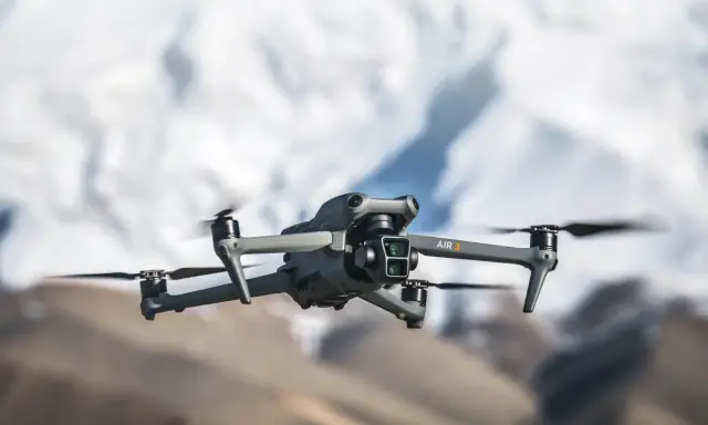 Szary dron DJI Air 3 z podwójną kamerą unosi się w powietrzu na tle rozmytych górskich szczytów.