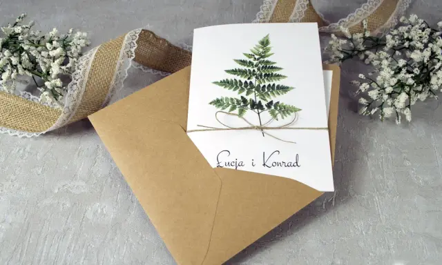 Eleganckie zaproszenie ślubne z paprocią i imionami pary. W zestawie koperta i ozdobna wstążka. To, co musi zawierać zaproszenie, to jego piękny design.