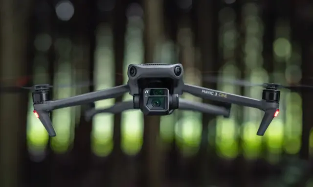 Szary dron Mavic 3 Cine zawieszony w powietrzu, z rozmytym zielonym lasem w tle.