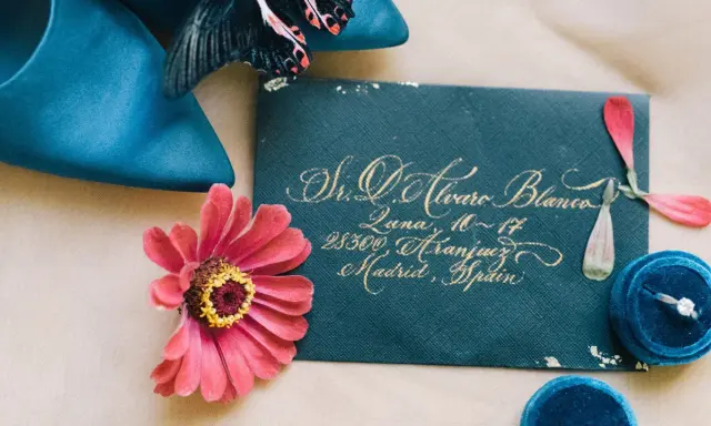Eleganckie zaproszenie na wesele 2 tygodnie przed, z wykaligrafowanym adresem, obok błękitnych butów, kwiatu i pudełka z pierścionkiem.