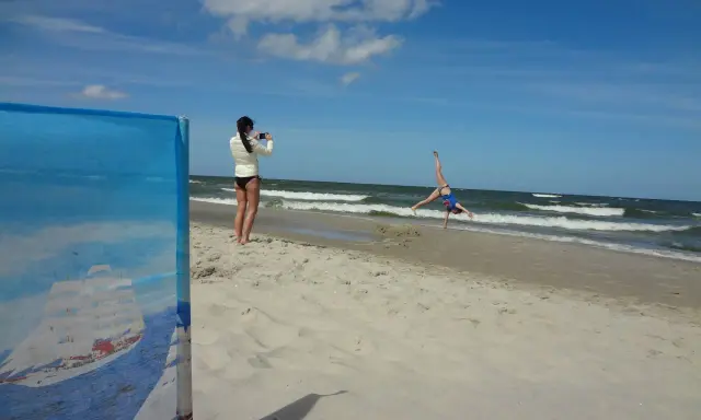 Dziewczyna robi fikołka na plaży, podczas gdy druga filmuje ją telefonem. Krzywy horyzont dodaje dynamiki scenie.