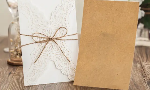Eleganckie, białe zaproszenie ślubne z koronkowym wzorem, przewiązane sznurkiem. Obok leży brązowa koperta, gotowa na ręczne wypisywanie zaproszeń ślubnych.