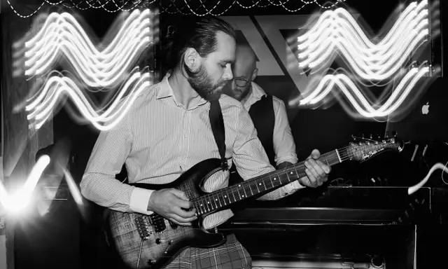 Muzyk gra na gitarze elektrycznej, a za nim stoi drugi mężczyzna. Efekt fal świetlnych sugeruje dynamiczną ekspozycję co to za moment.