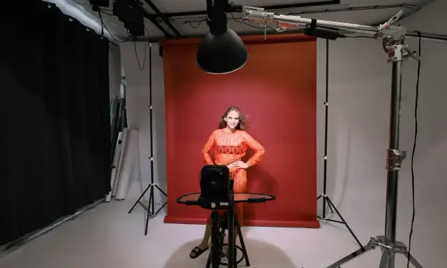 Oświetlenie studyjne przykłady: modelka w pomarańczowej sukience pozuje na tle czerwonego tła, otoczona sprzętem fotograficznym i lampami.
