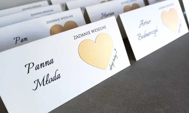 Białe karty z imionami gości i złotymi sercami, zapowiadające śmieszne zadania dla gości weselnych.