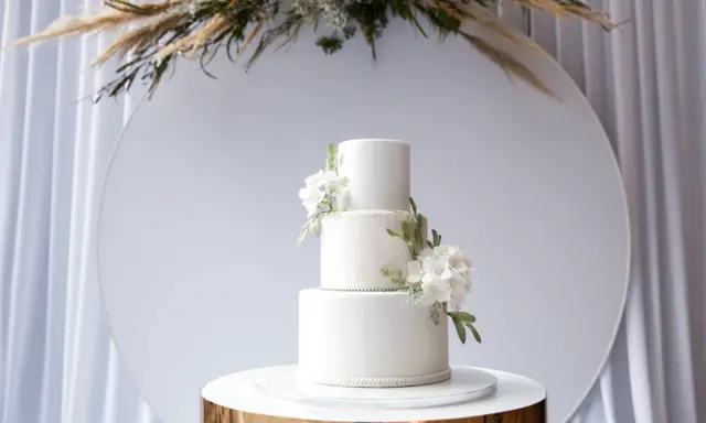 Elegancki, trójpoziomowy tort weselny w nowoczesnym stylu, ozdobiony białymi kwiatami i zielonymi liśćmi. Idealny na wyjątkowe okazje.