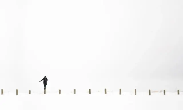 Osoba w czarnym płaszczu balansuje na słupku w śnieżnym krajobrazie. Minimalistyczna, high key fotografia.