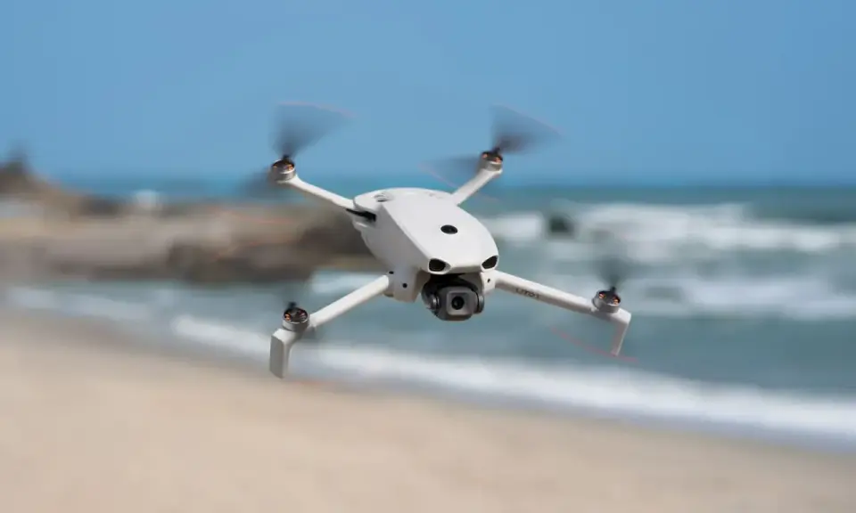 Biały dron DJI Lito X1 unosi się nad piaszczystą plażą, z morzem i niebem w tle.