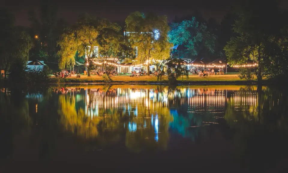 Nastrojowa fotografia nocna nad jeziorem, z oświetlonymi drzewami i budynkami, tworzącymi piękne odbicia w wodzie.