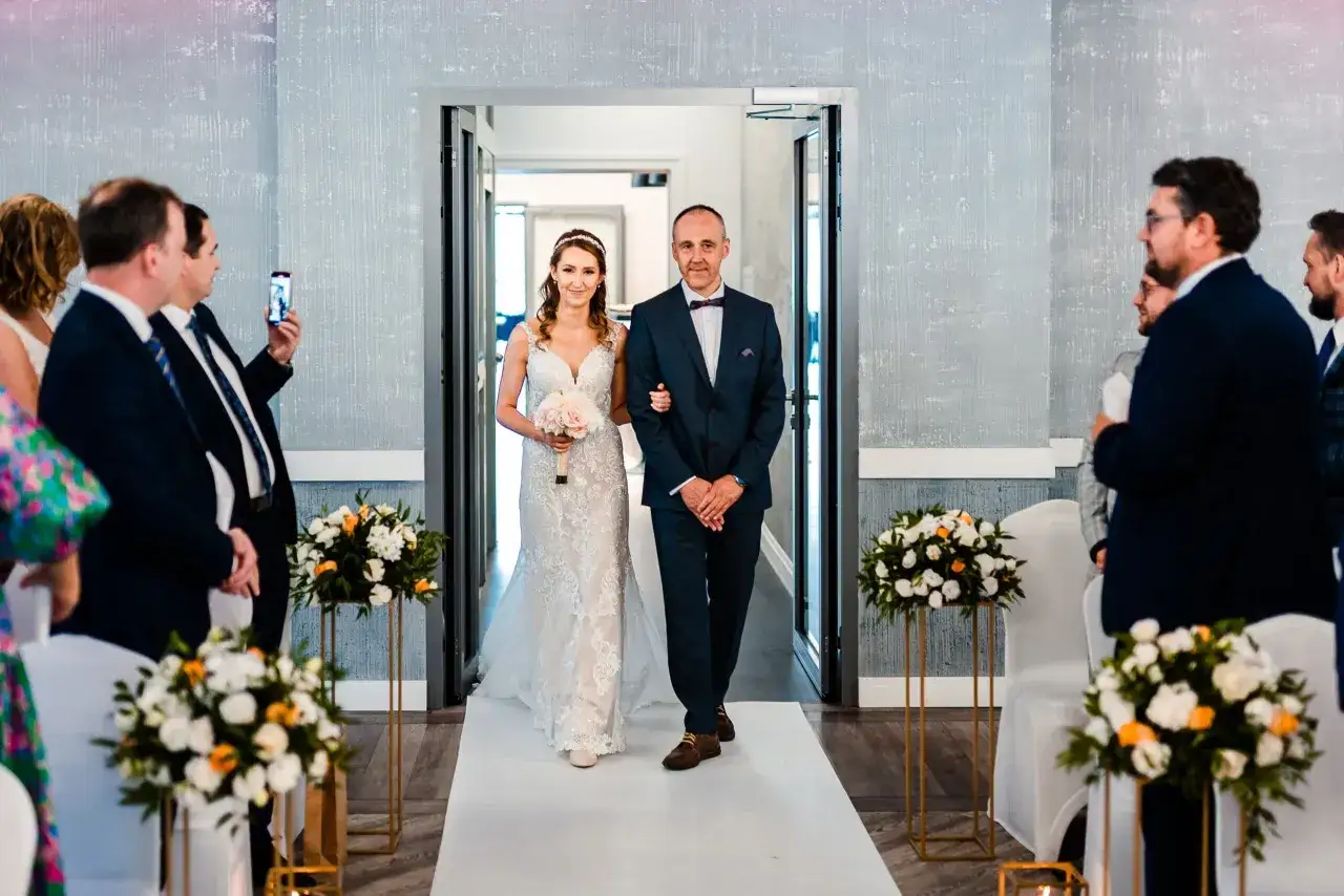 Panna młoda z ojcem idzie do ołtarza. Wśród gości, którzy mogą być świadkiem na ślubie, widać mężczyznę filmującego telefonem.