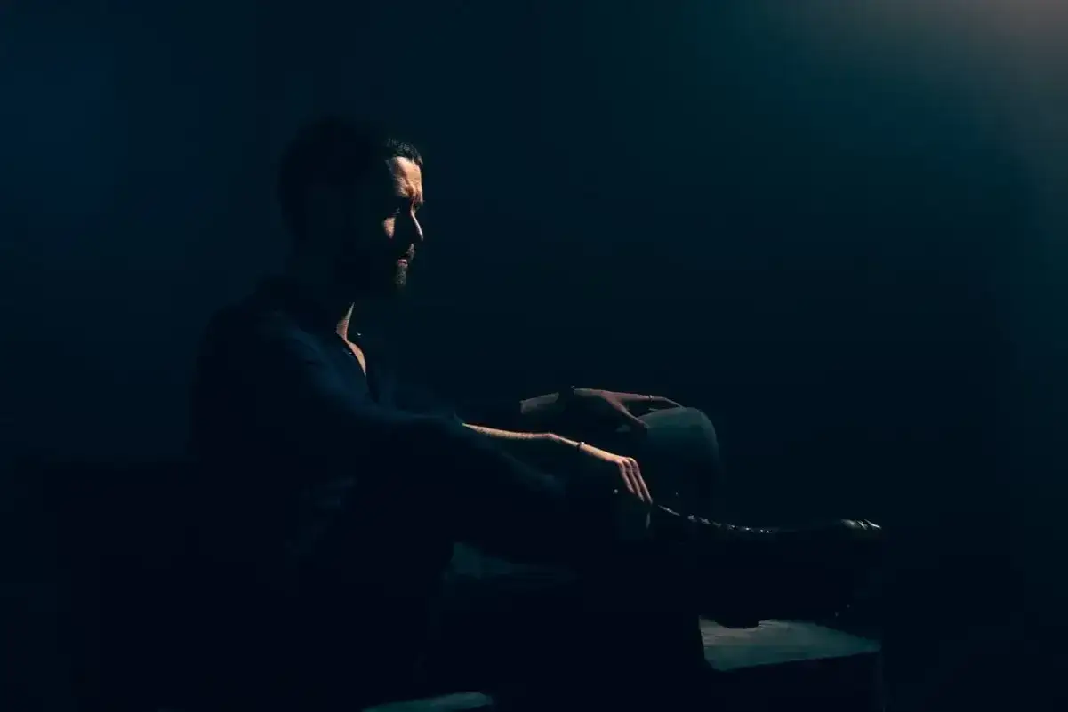 Mężczyzna w ciemnym pomieszczeniu, siedzi w profilu. Klimatyczna, low key fotografia podkreśla jego sylwetkę i skupienie.