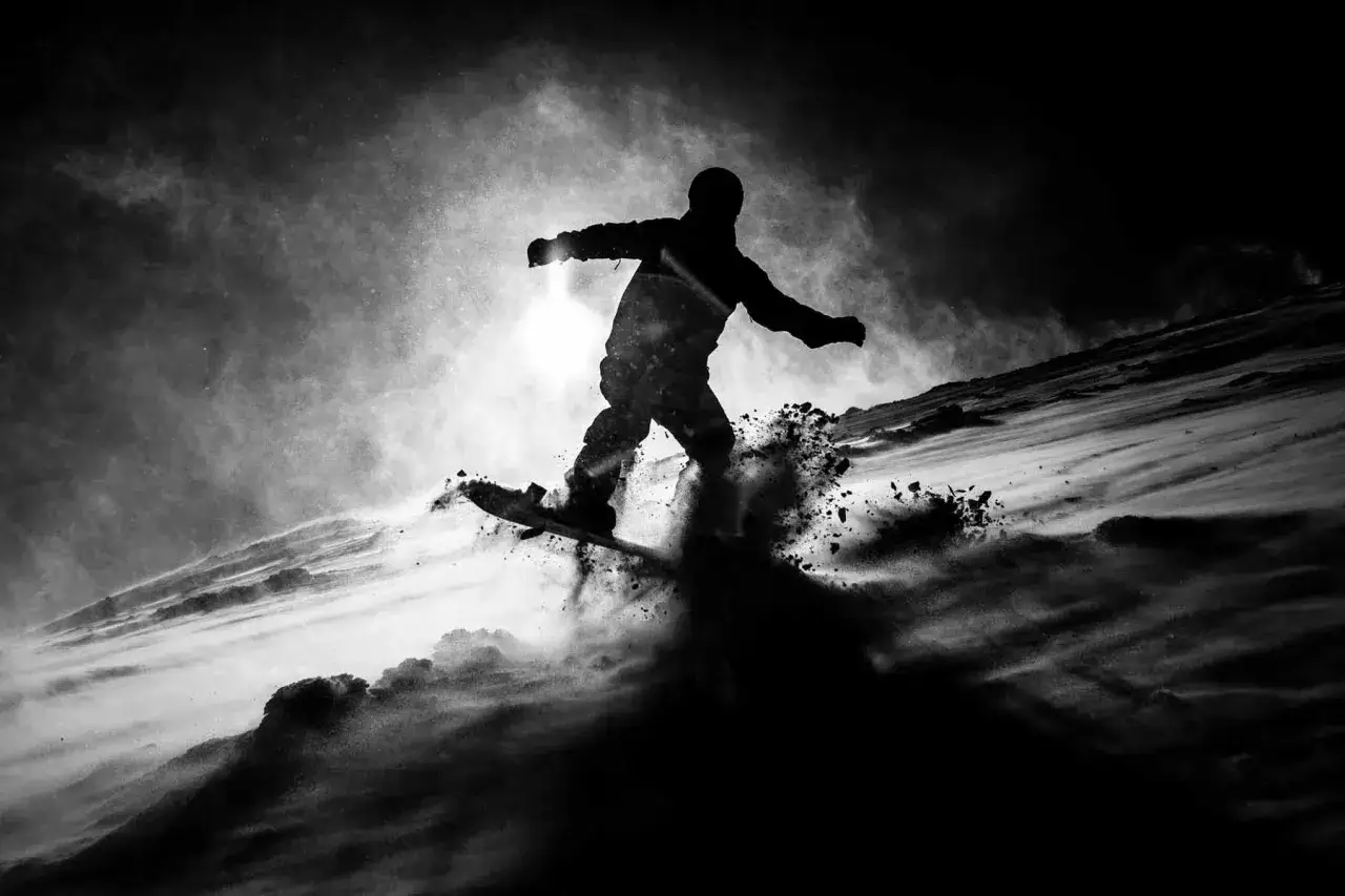 Silueta snowboardzisty w locie, z chmurami śniegu wokół. Dramatyczne światło i cień w fotografii podkreślają dynamikę ruchu.